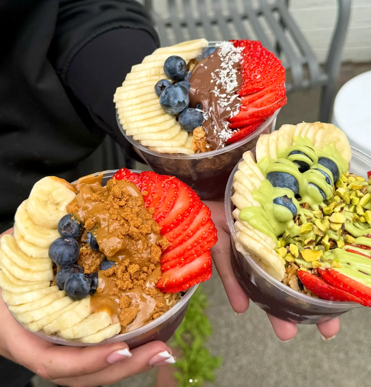 Three acai bowls topped with banana, berries, granola and nut butter held in hands