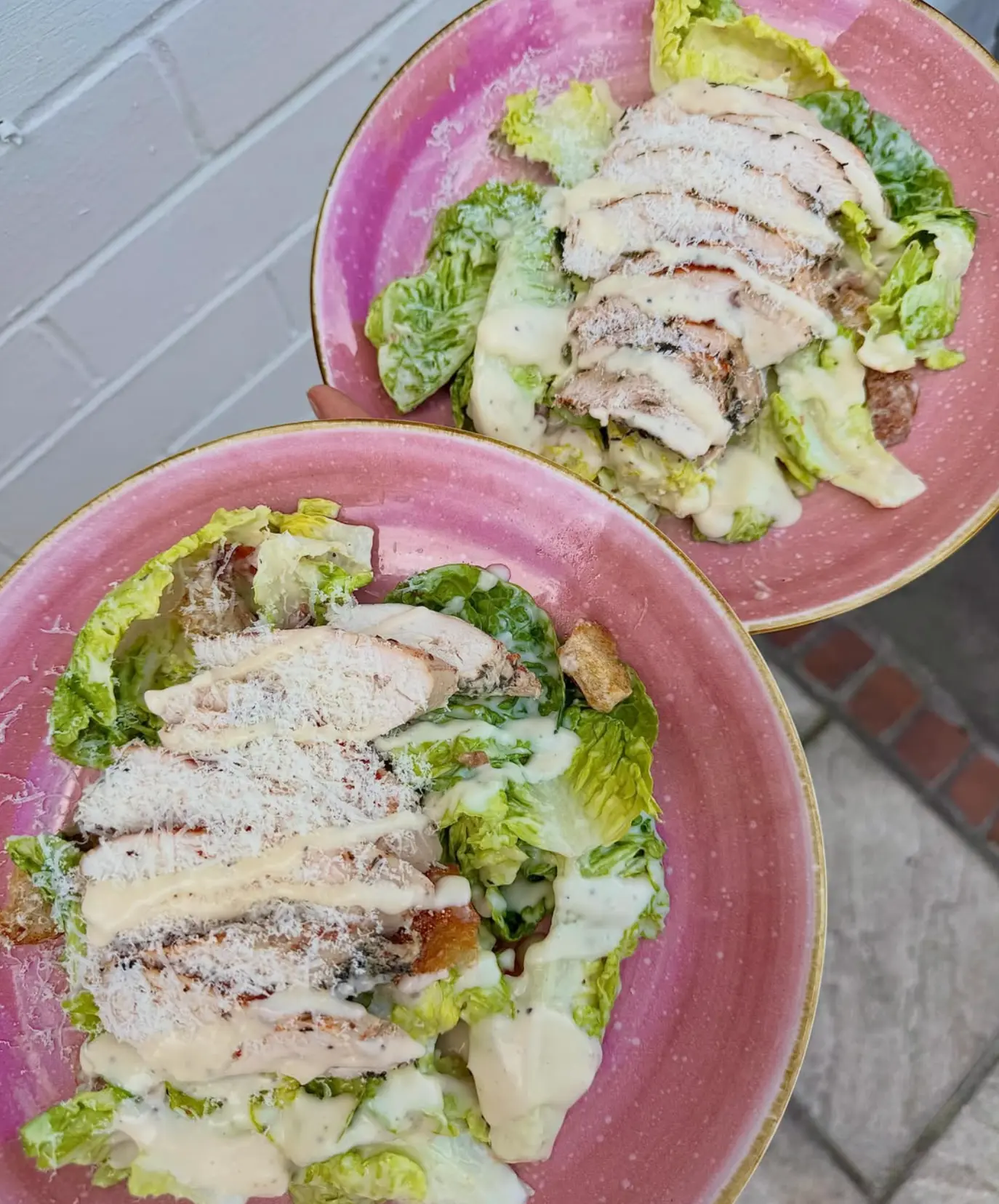 Two Caesar salads with sliced chicken and shaved parmesan on pink plates